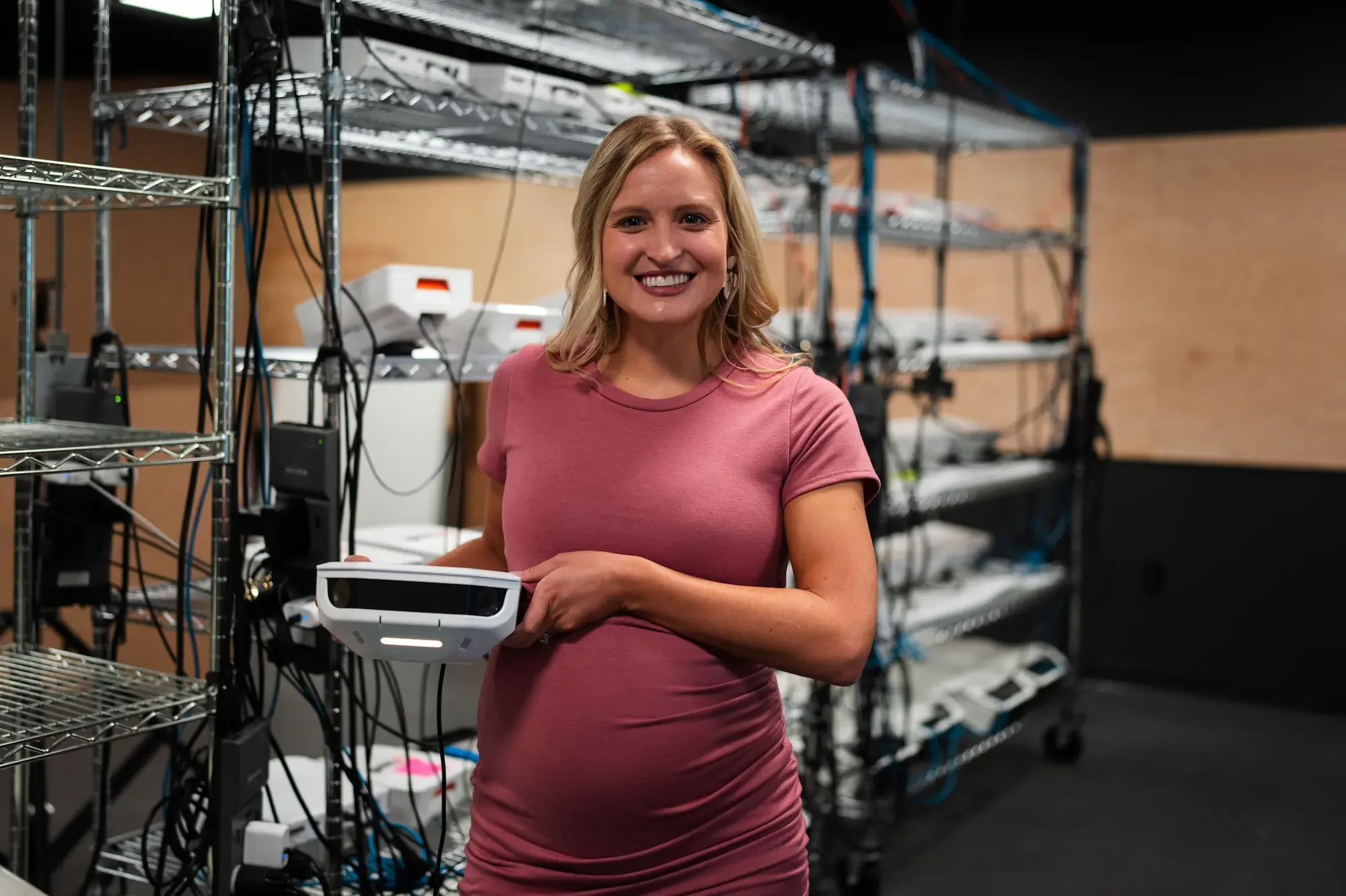 Photo of Tarryn Moss holding a Hudl Focus Outdoor camera. A shelf of cameras are behind her, for testing.
