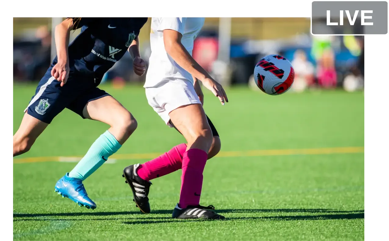 A close-up action shot of two soccer players competing for a ball on a green field, one in a navy kit with teal socks and another in a white kit with hot pink socks, featuring a 'LIVE' badge overlay in the upper right corner.