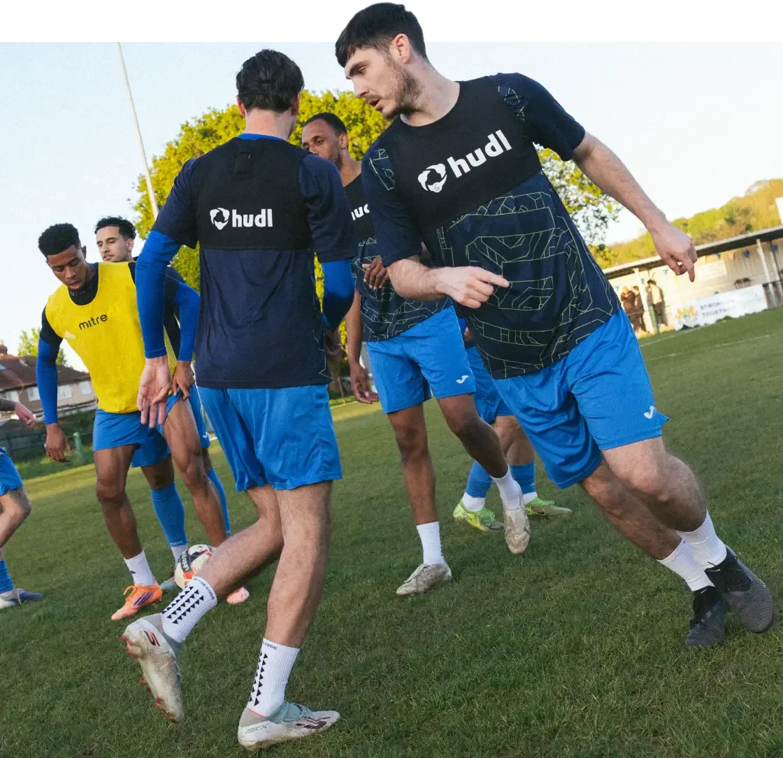 A group of soccer players training on a grass field, with two prominent players in the foreground wearing blue shorts and black Hudl performance tracking vests over their training shirts.