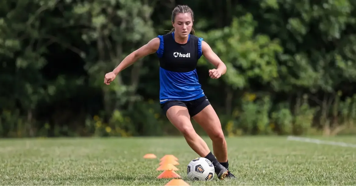 A female soccer player in a blue training top and a black Hudl-branded wearable tech vest dribbling a soccer ball through orange cones on a grass field.