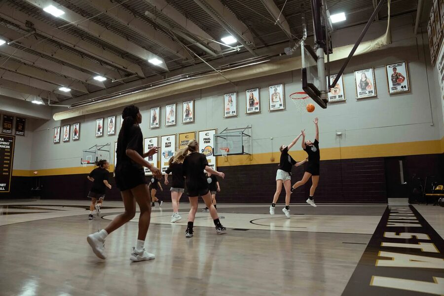 A wide shot shows a group of female basketball players practicing in a well-lit gymnasium. The players are dressed in black athletic wear, and two are actively jumping for a rebound under the hoop, one reaching towards the orange basketball. The gym features a light-colored court with "EMPORIA STATE" written along the baseline in black lettering with a yellow outline. The walls are light grey with a prominent black and yellow stripe running along the lower section. Numerous framed photos and banners, likely commemorating past athletes or achievements, adorn the walls. The ceiling has visible beams and bright fluorescent lighting.