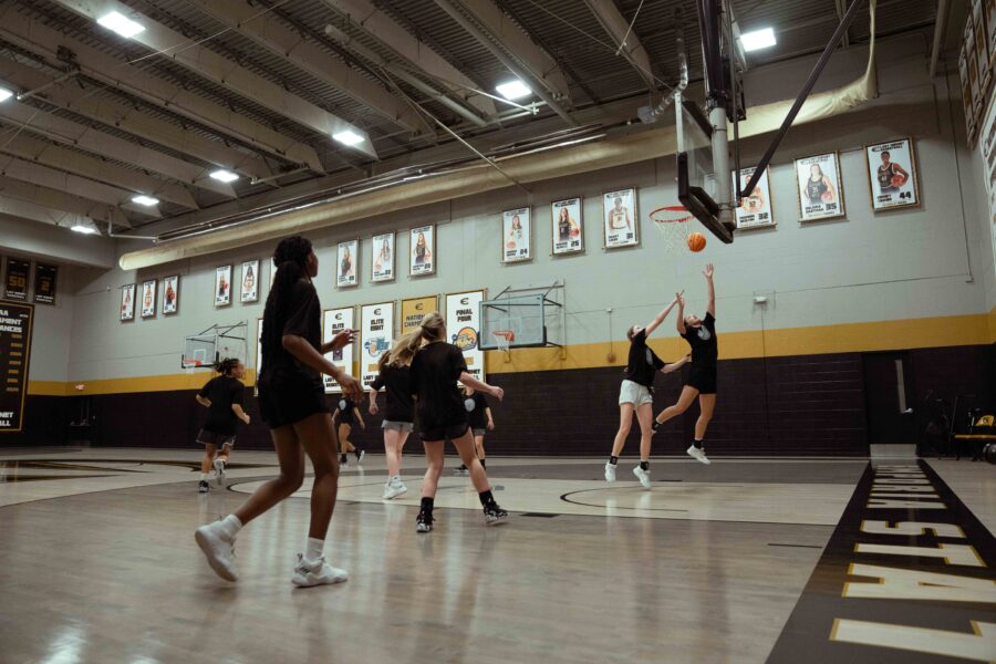 A wide shot shows a group of female basketball players practicing in a well-lit gymnasium. The players are dressed in black athletic wear, and two are actively jumping for a rebound under the hoop, one reaching towards the orange basketball. The gym features a light-colored court with "EMPORIA STATE" written along the baseline in black lettering with a yellow outline. The walls are light grey with a prominent black and yellow stripe running along the lower section. Numerous framed photos and banners, likely commemorating past athletes or achievements, adorn the walls. The ceiling has visible beams and bright fluorescent lighting.