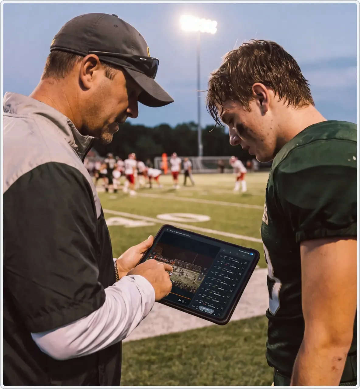 A football coach on the sideline of a live game, reviewing film with a player.
