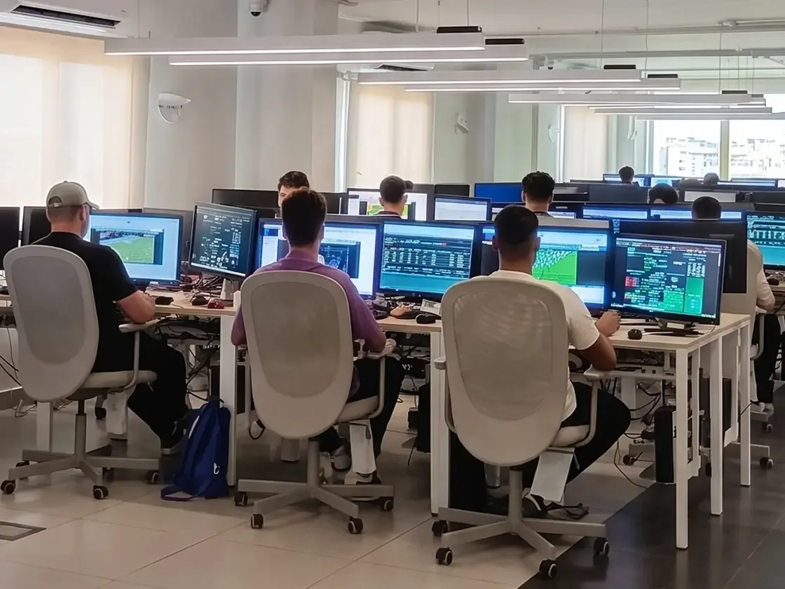 A bright, open-plan office space with multiple analysts sitting at rows of desks with dual-monitor setups, reviewing sports match footage and data analytics software.