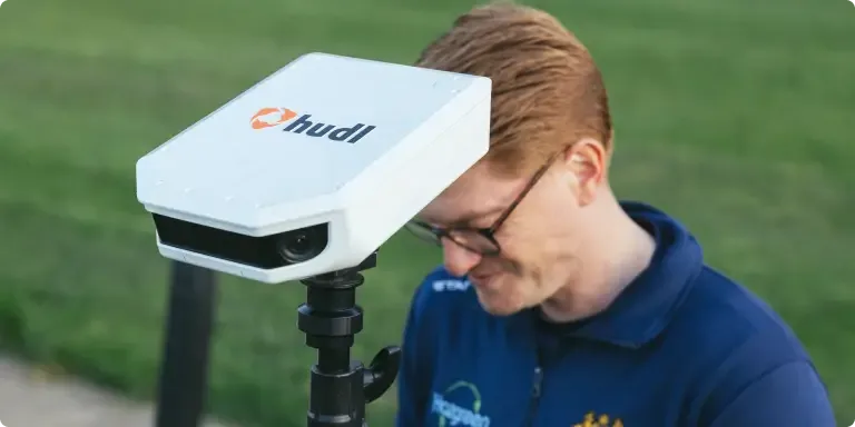 A Hudl Focus Flex smart camera unit mounted on a tripod in the foreground, with a man wearing glasses and a navy jacket standing behind it on a grass field.