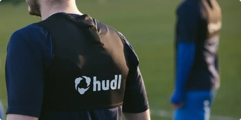 A close-up shot from behind an athlete wearing a black Hudl-branded athletic tracking vest on a grass training field, with another player blurred in the background.
