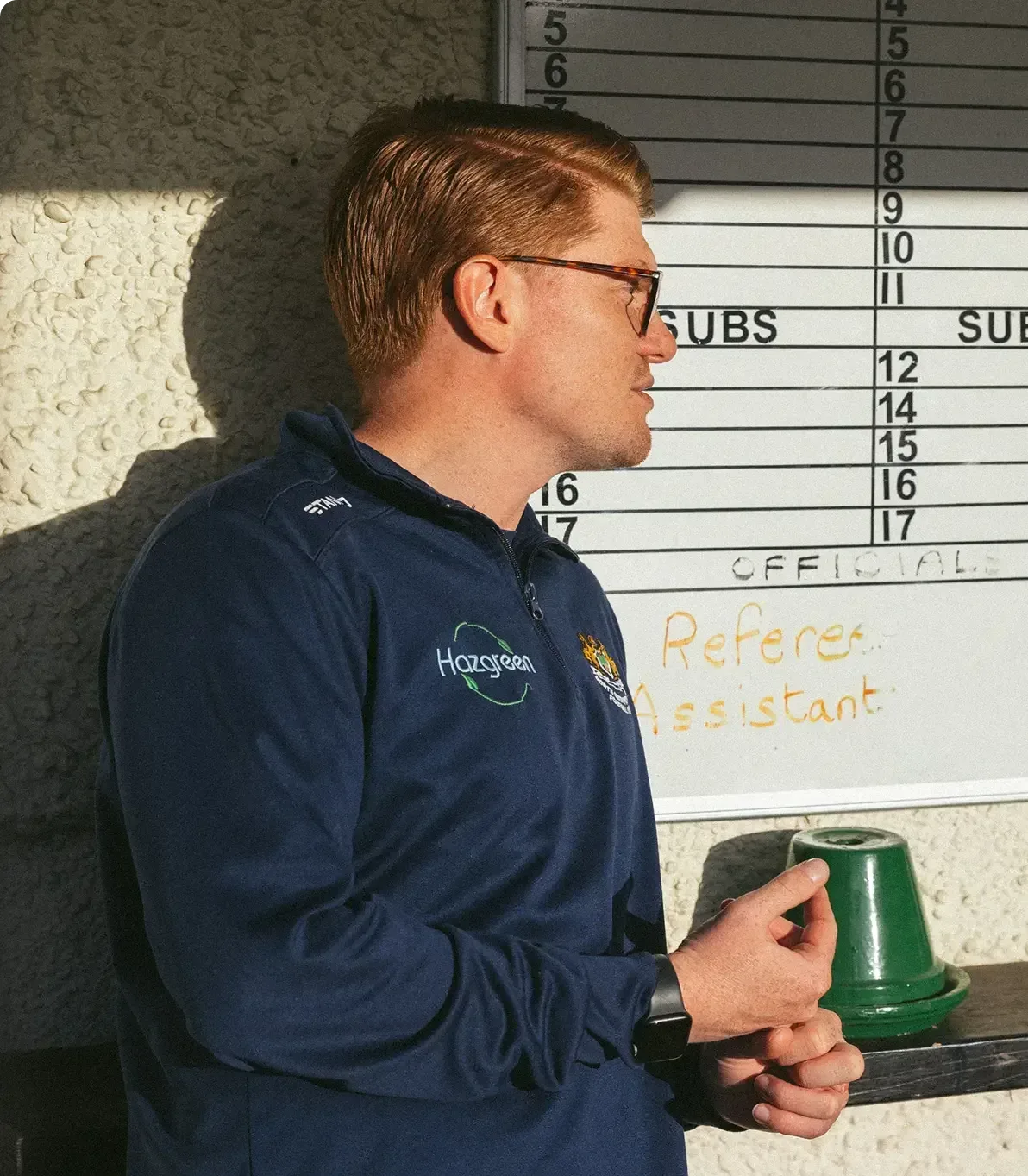 Football Club Manager Joey Gritt leaning against a wall with a whiteboard, looking away.