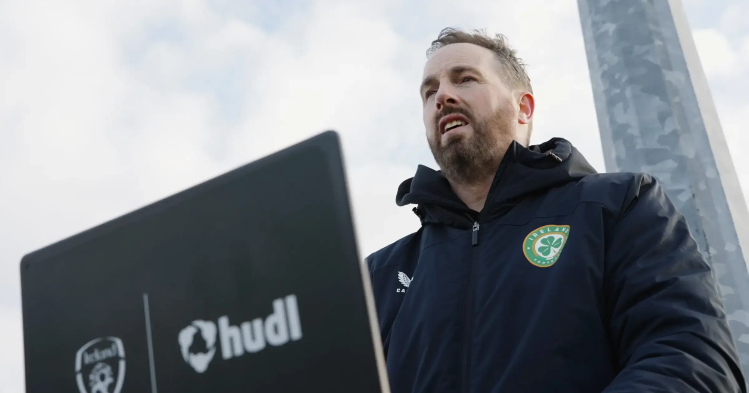 A man in an Ireland national team jacket standing outdoors behind a laptop featuring the Ireland and Hudl logos.