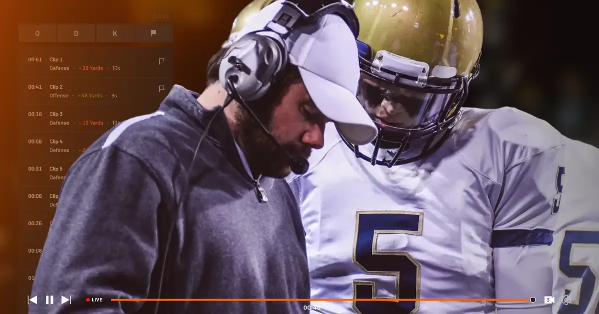 A football coach wearing a headset and white cap reviews game clips on a digital sideline replay screen with a player in a gold helmet.