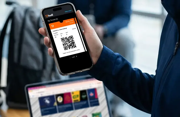 A person holding a smartphone that displays a digital football game ticket with a QR code and student seating information. In the background, a laptop screen and a backpack are partially visible.